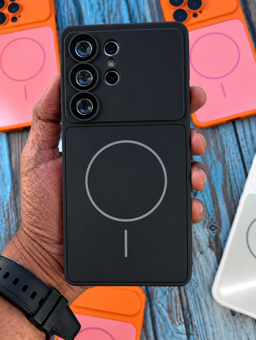 A hand holds a black magnetic phone case with a split dual-camera cutout, shown against a blue wooden background with other colorful phone cases.