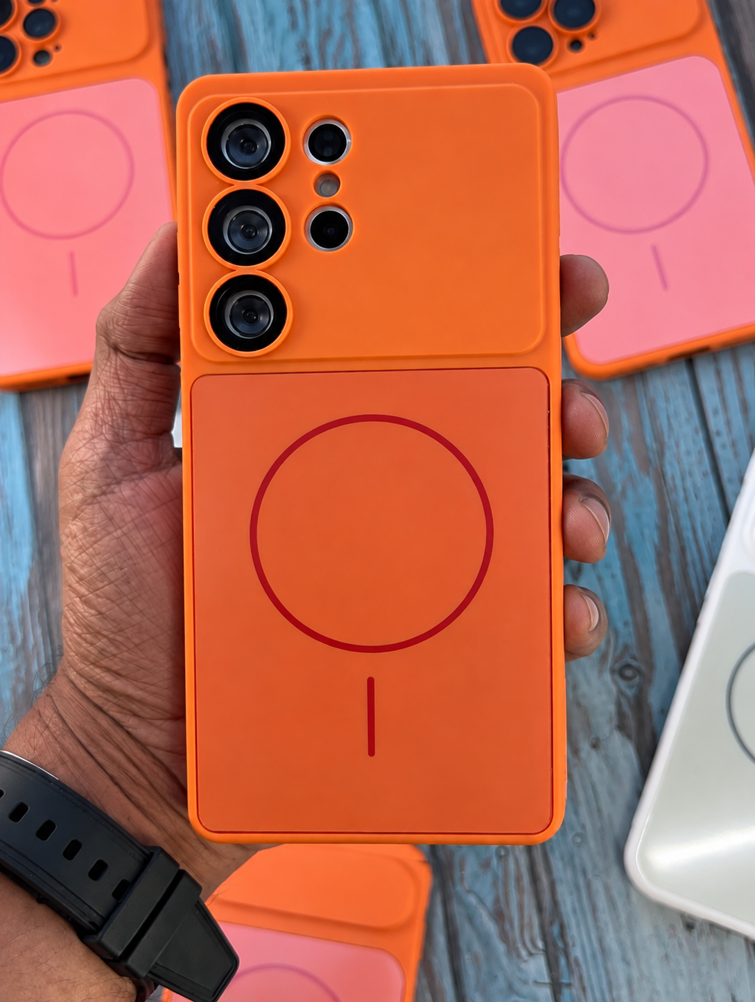 An orange magnetic phone case with a fold-out power bank design, held in a hand over a blue wooden surface with other phone cases in the background.