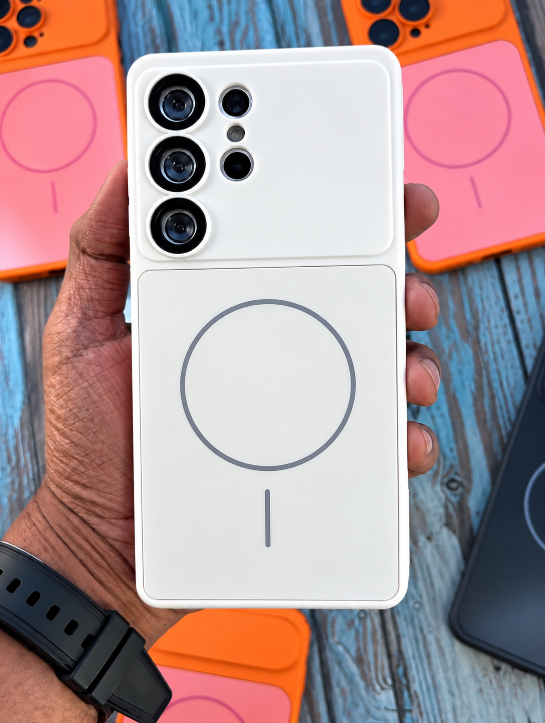 A hand holds a white MagSafe-compatible phone case designed for a Samsung Galaxy Ultra smartphone, showing the magnetic ring design on the back. Other colorful phone cases are visible in the background on a rustic blue wooden surface.