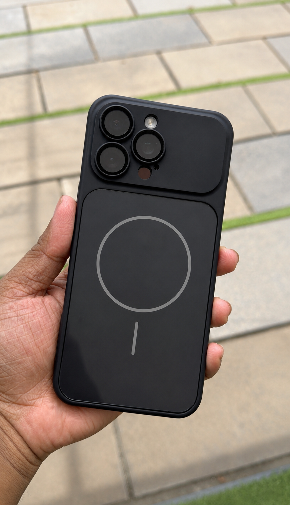 A hand holding a black magnetic phone case with a built-in camera cutout and a large MagSafe-compatible ring on the back, photographed outdoors against a blurred paved background.
