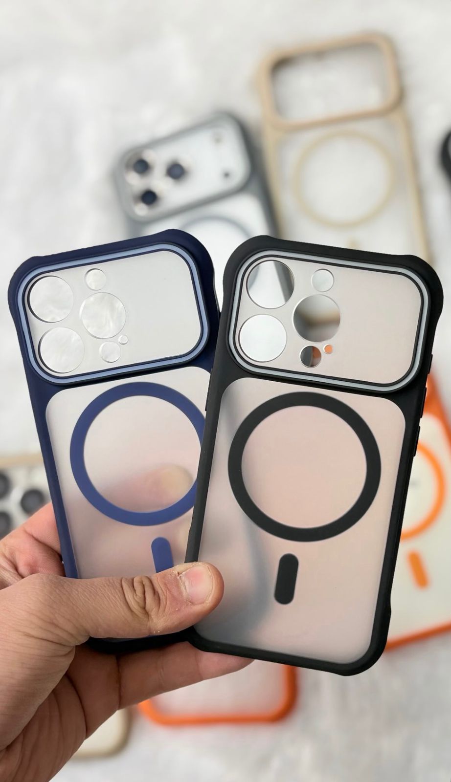 Two MagSafe-compatible phone cases held in a hand, one in navy blue and one in black, with more cases blurred in the background.
