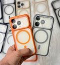 A hand holds two MagSafe-compatible phone cases in front of a blurred background of more cases. The case on the left is bright orange and white, and the case on the right is dark gray and white.
