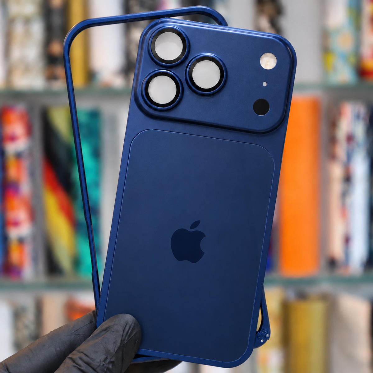 A close-up of a hand wearing a black glove holding a blue protective phone case with a matching blue iPhone. The case features a raised camera module and a large rectangular magnetic card slot on the back, set against a blurred background of bookshelves.