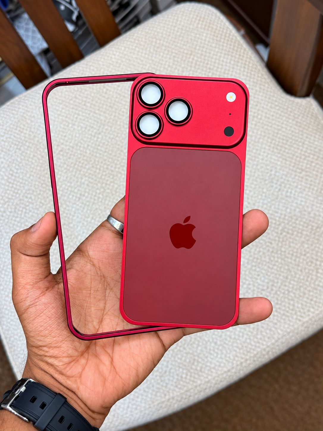 A hand holds a red iPhone case with a matching red back plate and clear outer bumper over an iPhone, with a beige upholstered chair and wooden chair legs in the background.