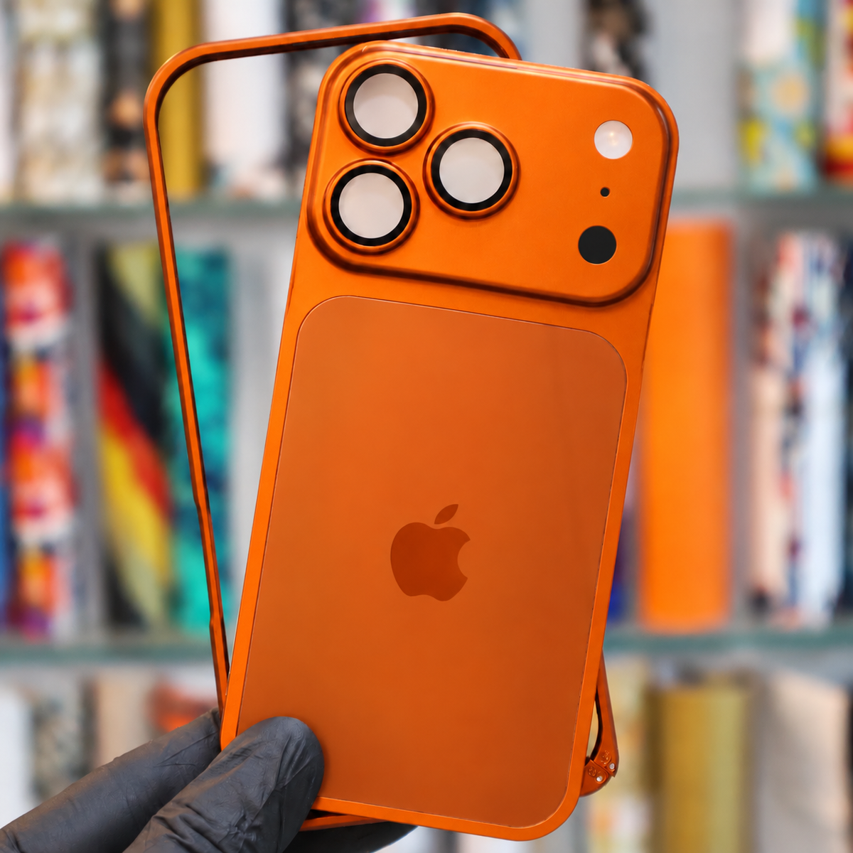 An orange protective phone case with a built-in MagSafe magnetic ring, shown fitted on a smartphone held by a gloved hand in front of a blurred bookshelf background.