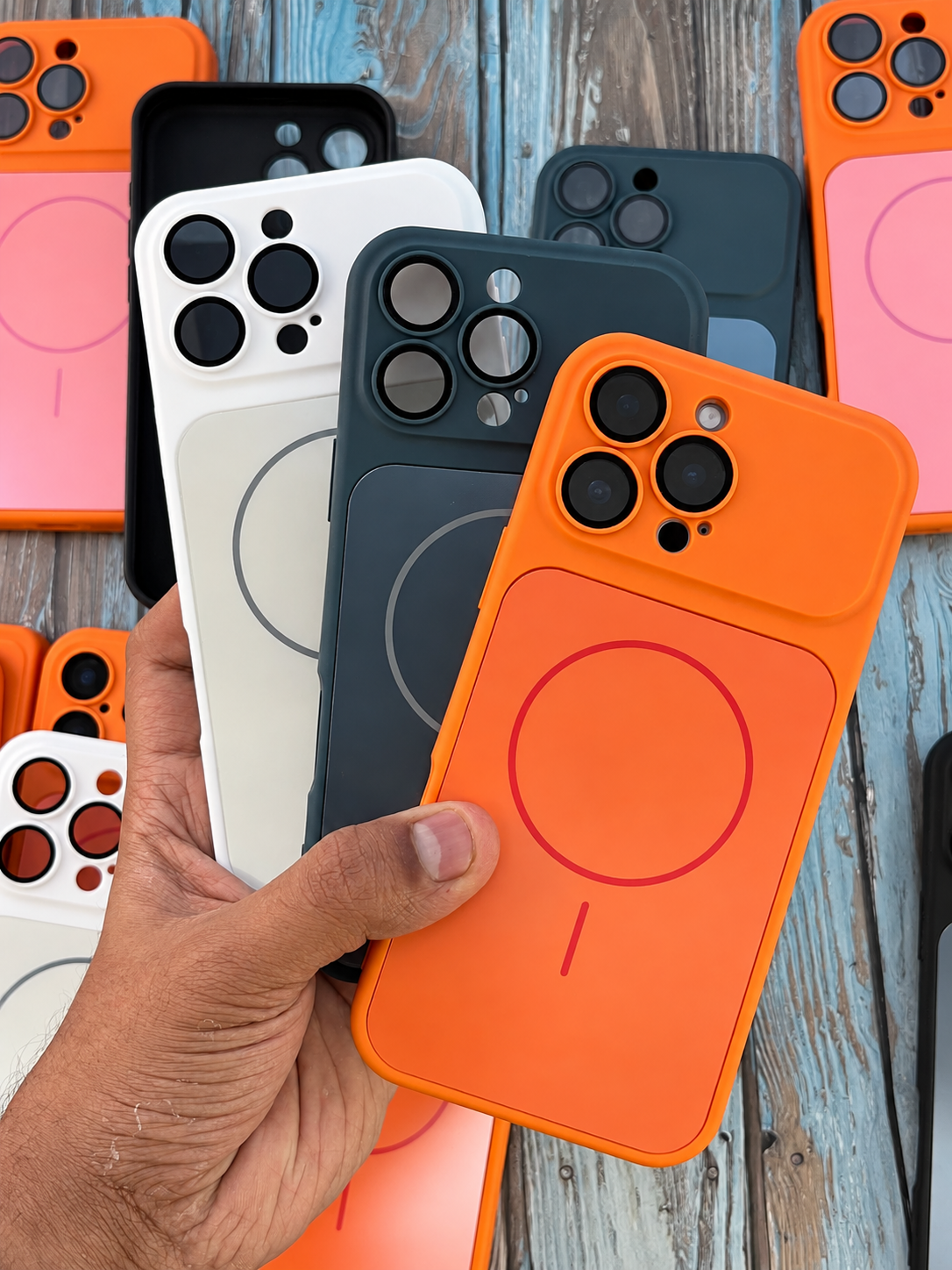 A hand holds a stack of MagSafe-compatible phone cases in various colors, including orange, white, black, and dark gray, against a rustic blue wooden background.