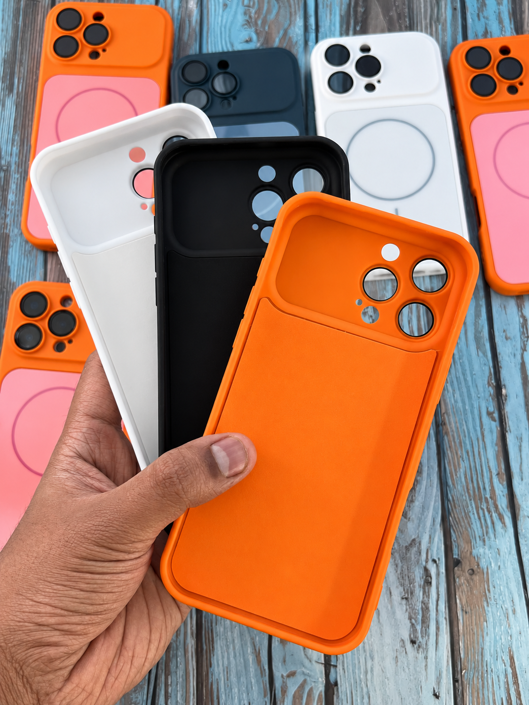 A hand holds an orange phone case in the foreground, with several other phone cases in white, black, pink, and orange arranged on a rustic blue wooden surface.