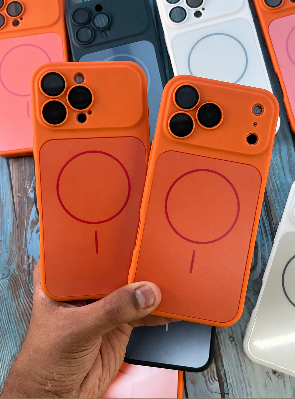 Two bright orange MagSafe-compatible phone cases are held in a hand over a rustic wooden surface, with other colored phone cases visible in the background.