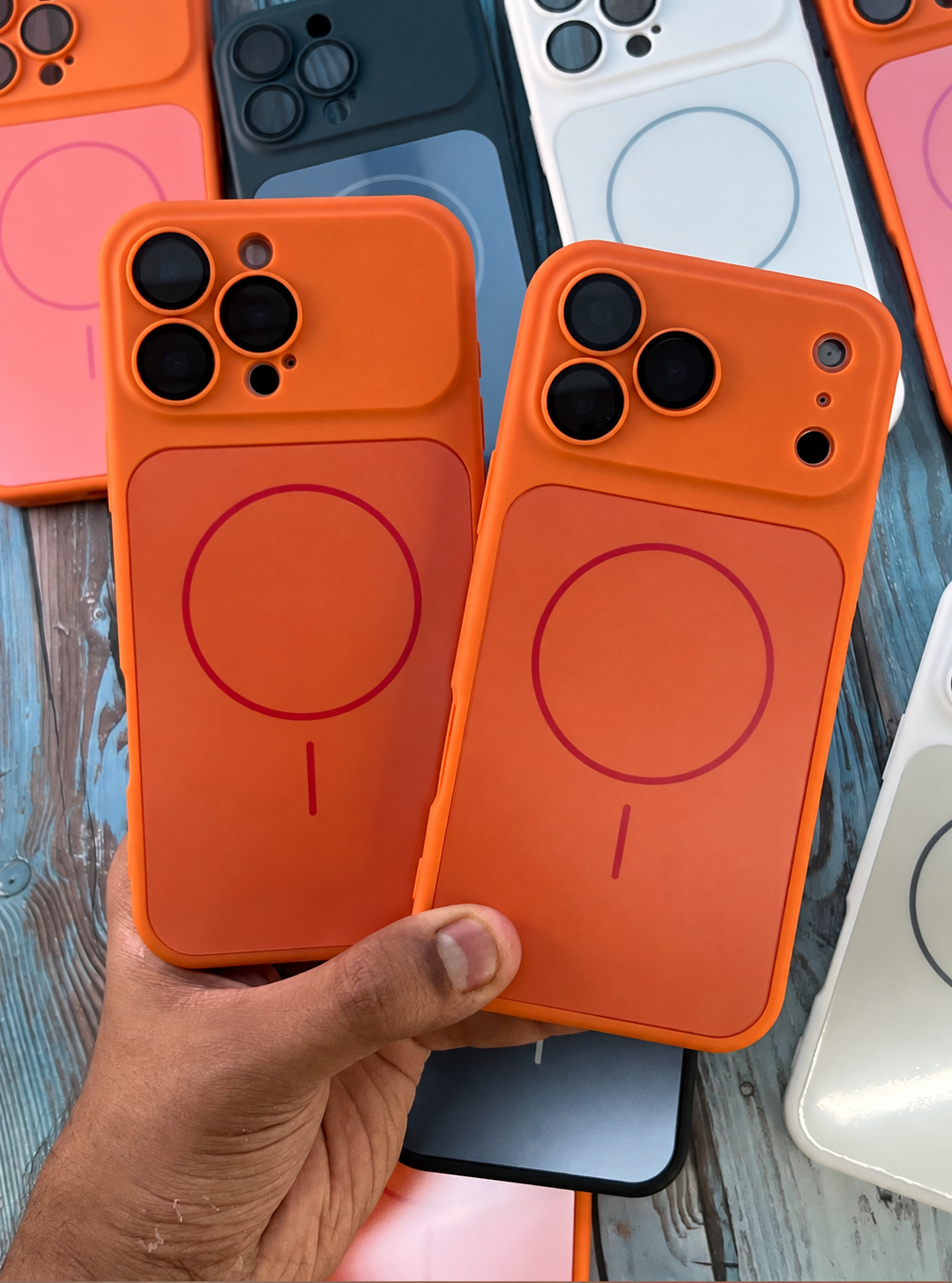 Two bright orange MagSafe-compatible phone cases are held in a hand over a rustic wooden surface, with other colored phone cases visible in the background.