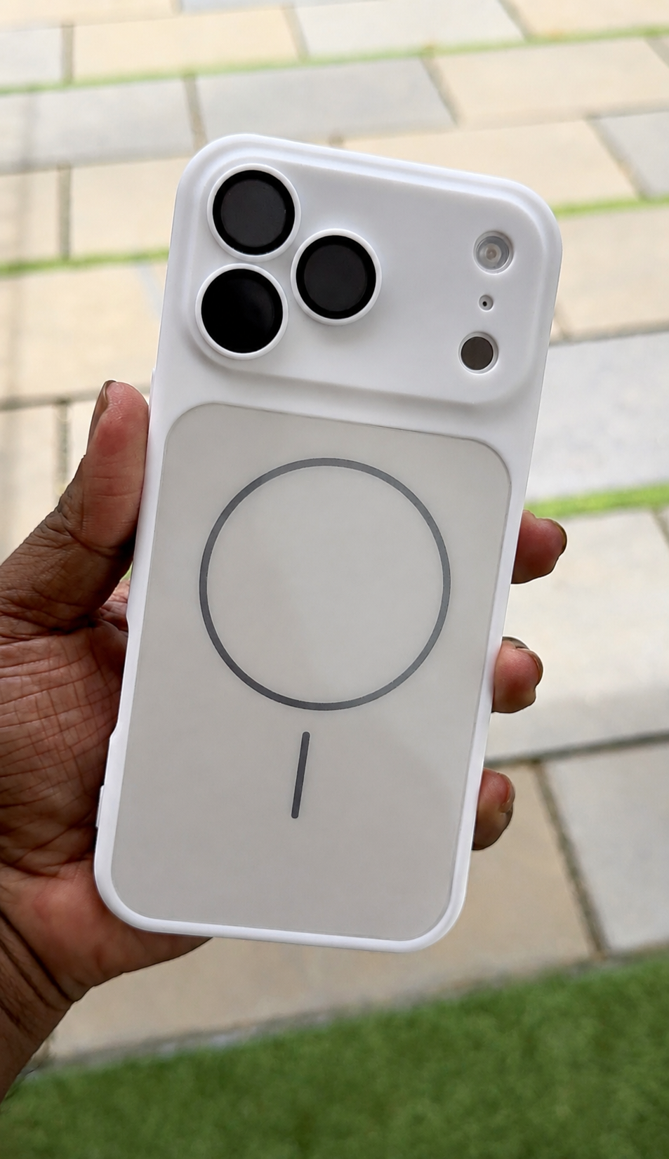 A hand holding a white MagSafe-compatible phone case outdoors on a paved walkway with grass visible in the background.